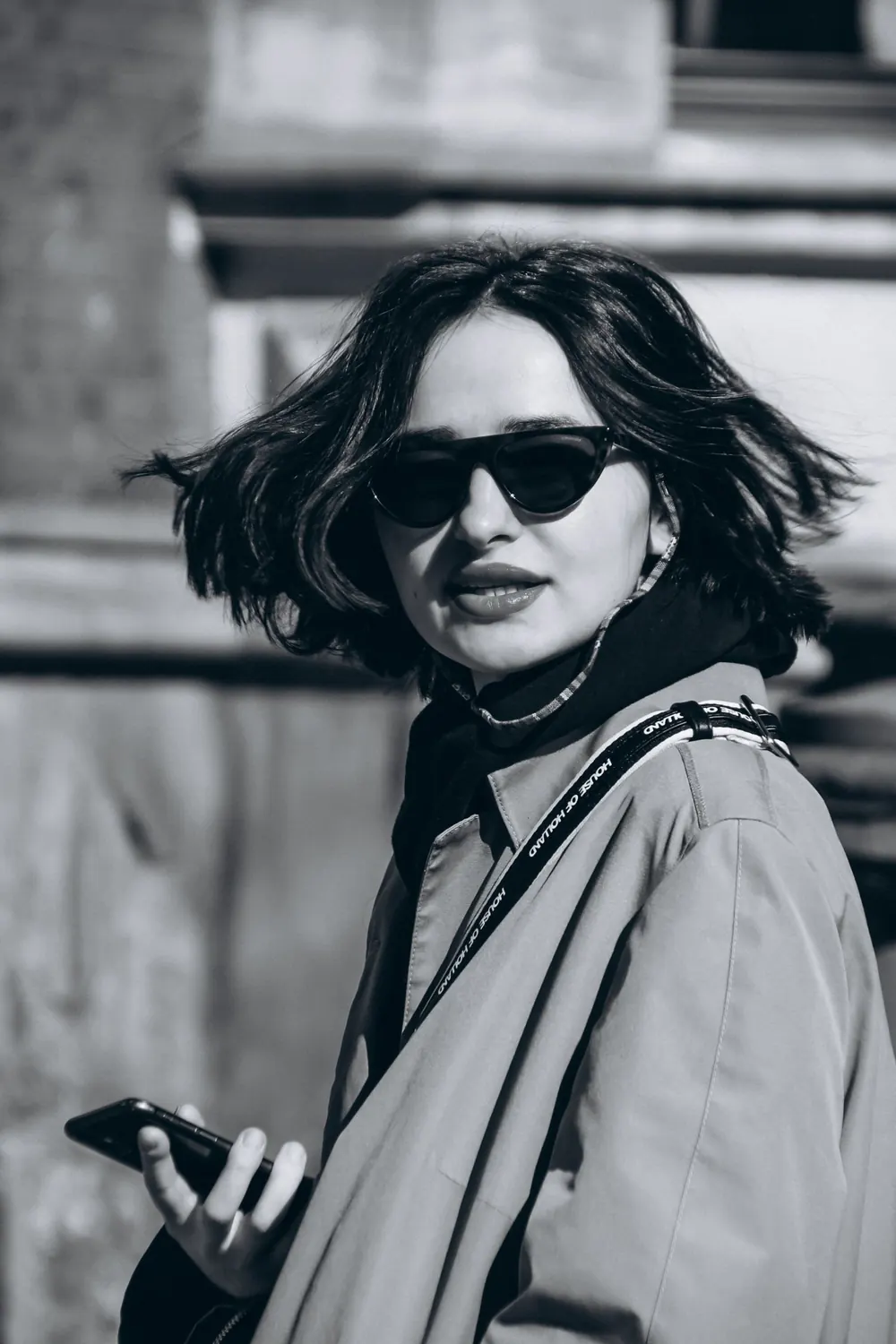 Fashionable woman with bob haircut and sunglasses holding a phone in a city setting.