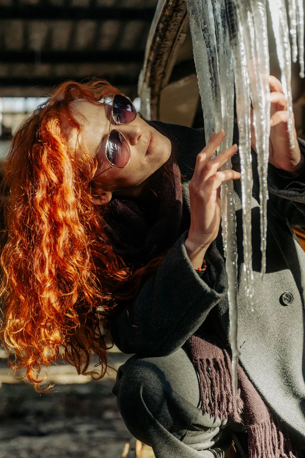 Red-haired woman in sunglasses touching icicles in sunlight, creating winter vibes.
