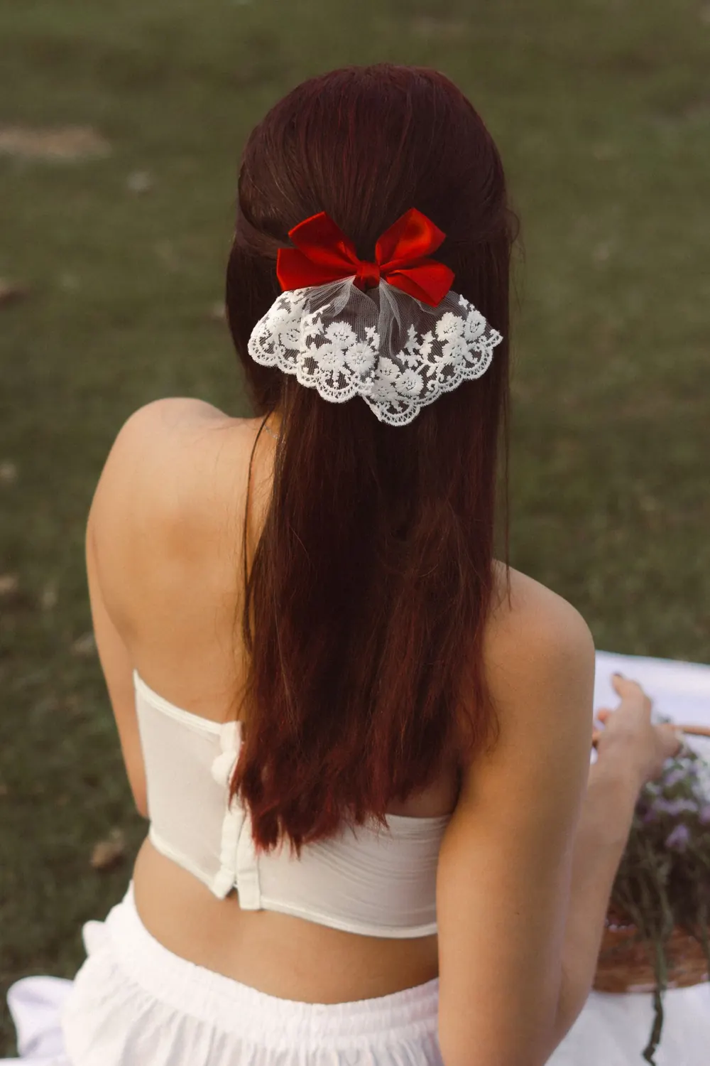 A woman with long hair adorned with a red ribbon and lace bow sits outdoors.