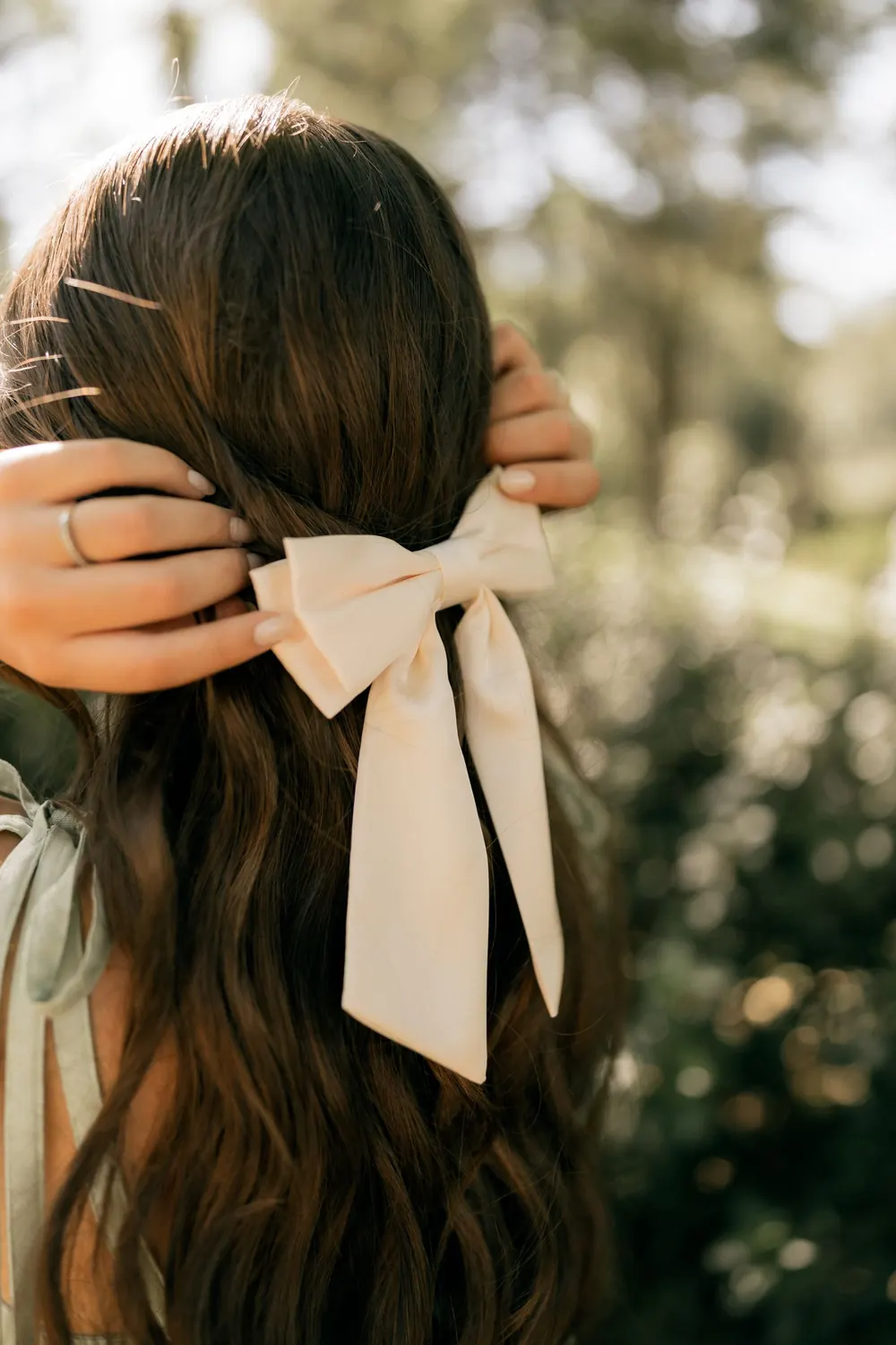 A woman styles her hair with a cream bow in a sunlit outdoor setting, evoking elegance.