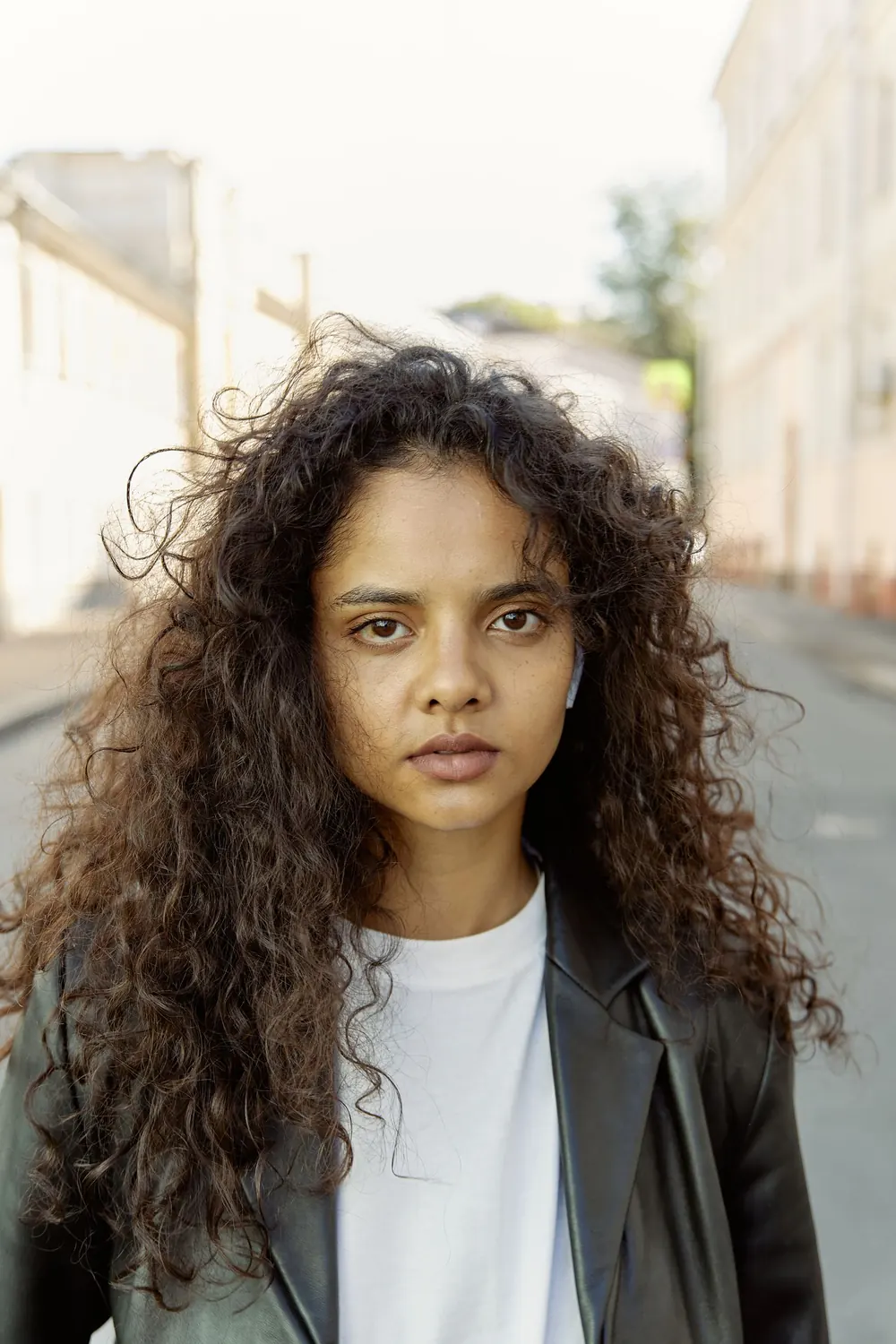 A woman with curly hair in a leather jacket stands confidently on a quiet street.