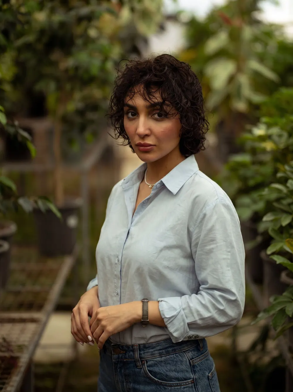 Woman with curly hair in a denim outfit poses confidently in a lush greenhouse.