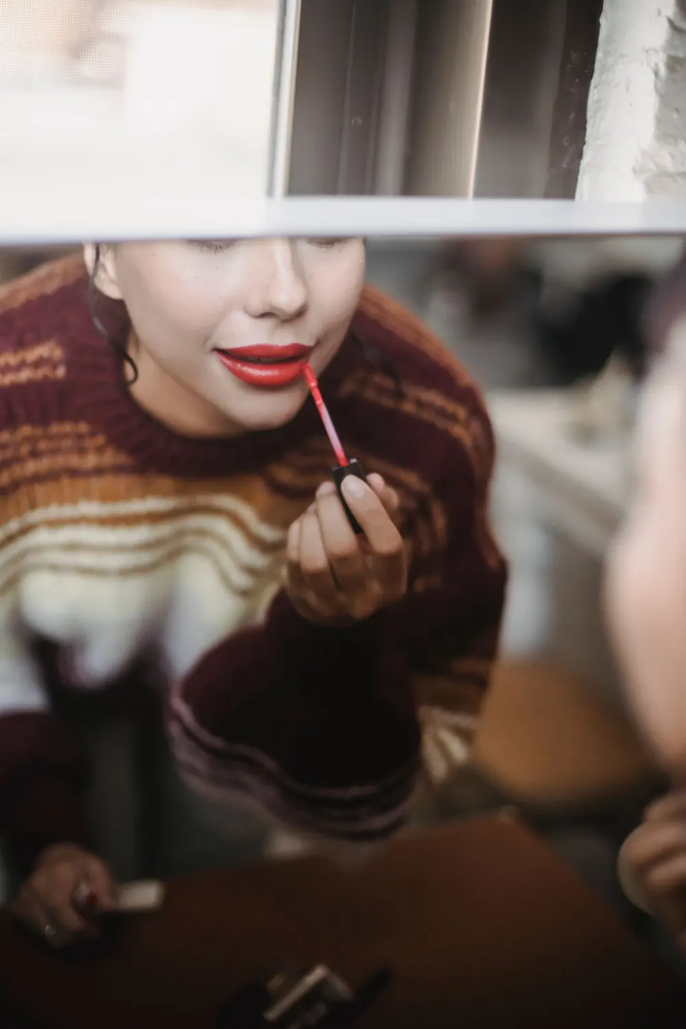 Young woman applying red lipstick in front of a mirror indoors.